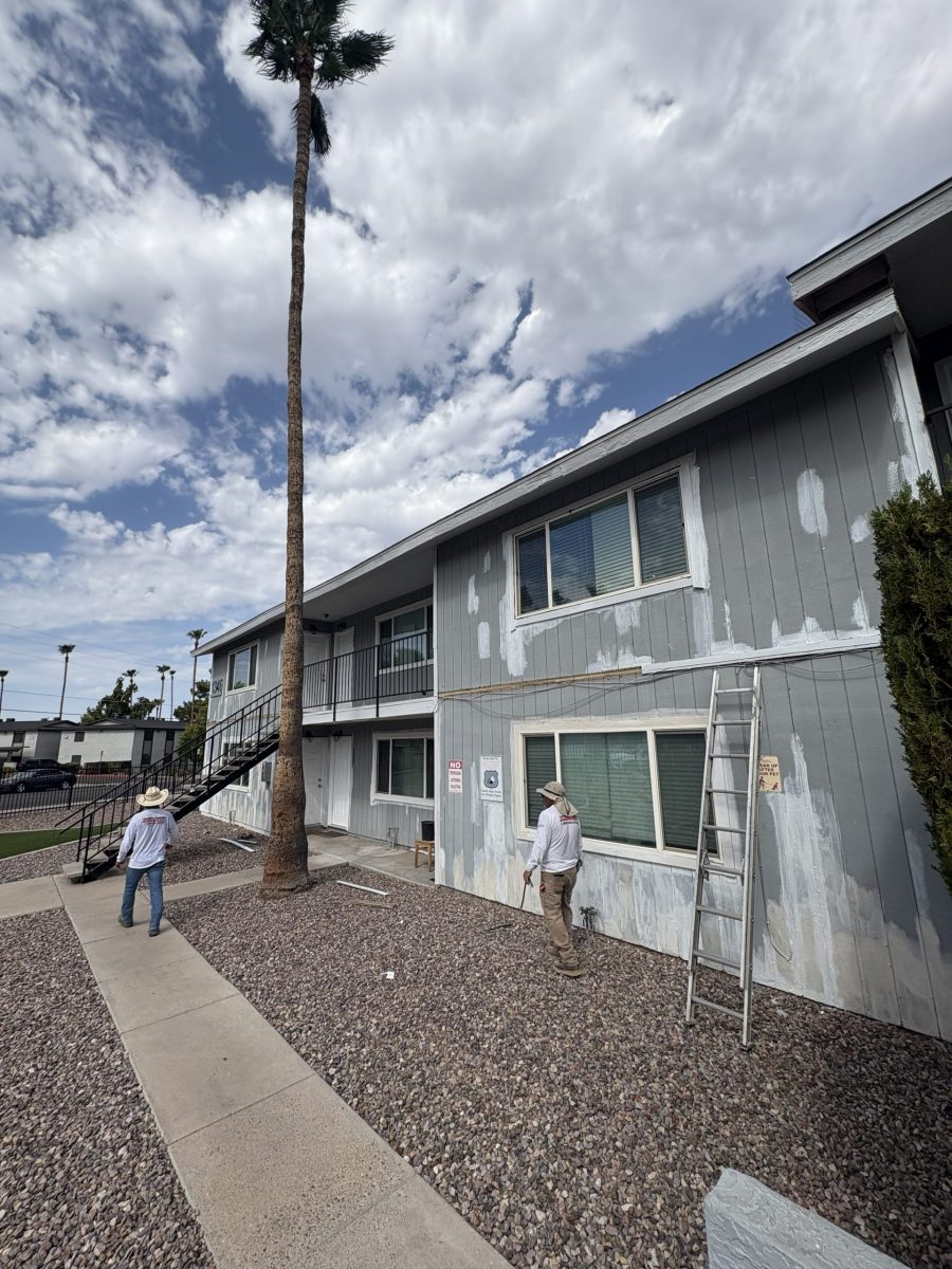 Before photo of a two-story Phoenix apartment building with faded blue-gray vertical siding showing patched and spot-primed areas, a ladder against the wall and a tall palm tree in front Preview Image 1