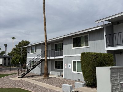 After photo of the same two-story Phoenix apartment building freshly painted light gray with white trim and black stair railings, a tall palm tree and gravel landscaping in front