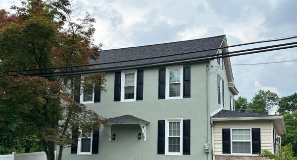 Historic-style Pennsylvania home with pale gray-green lap siding, black shutters, white bracketed entryway, and unpainted fieldstone wing.