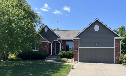 Kechi ranch home featuring charcoal gray siding, white arched entry trim, a gray garage door, and red brick skirt.