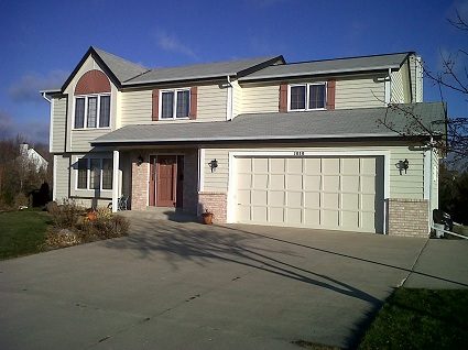 Two-story Waukesha home displaying light sage green siding, white trim, deep red shutters, a red front door, and a white garage door.