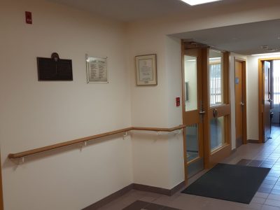Warm off-white commercial corridor in Stoney Creek featuring a wall-mounted wood handrail, brown tile flooring, and glass doors.