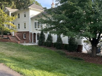 After photo of a two-story Colonial-style home in Lorton, Virginia with freshly painted cream lap siding, white trim, a red brick foundation and front steps, a cupola on the roof, and mature trees on a sloped lawn
